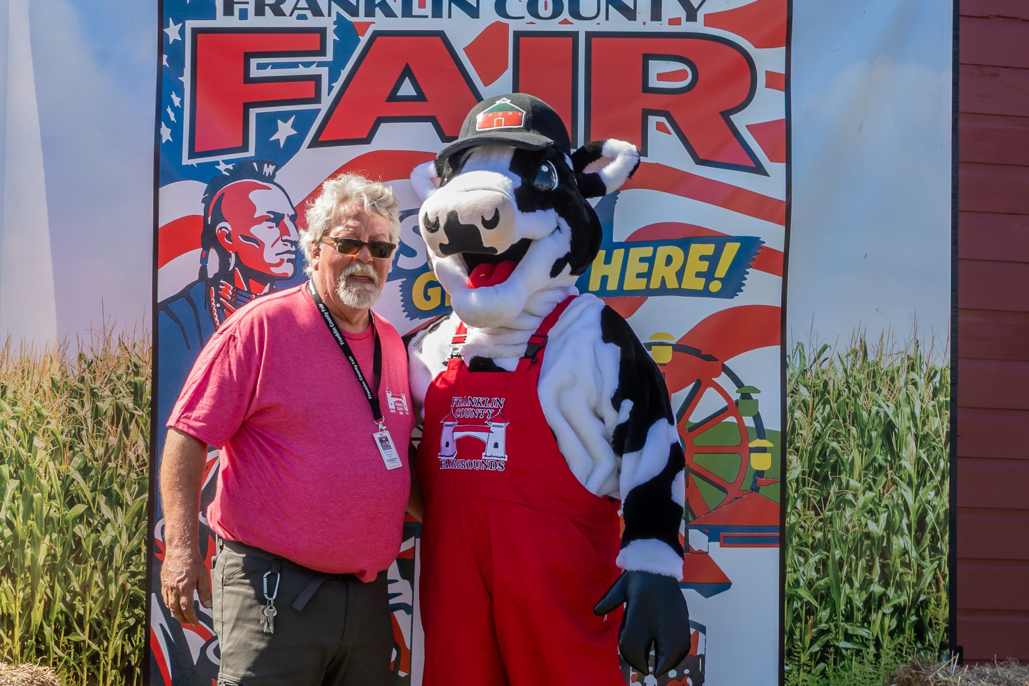 Steve DeJoy standing beside the cow mascot in front of the fair banner